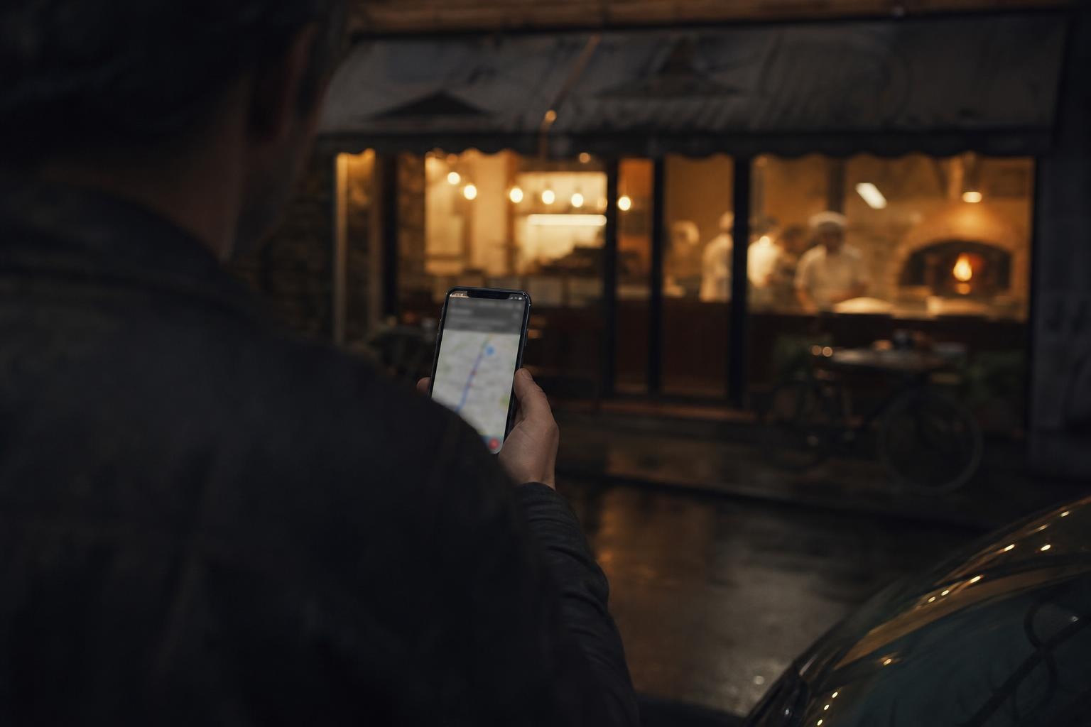 A hand holding a phone with a glowing map, a pizzeria storefront across the street